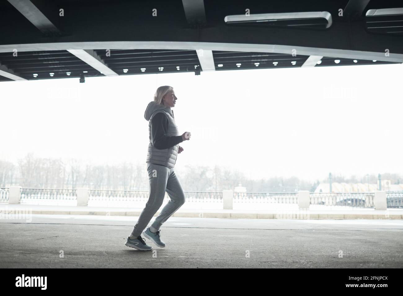 Fit Senior Frau in grauen Sportanzug Joggen allein unter Überbrücken Sie beim Training im Freien Stockfoto