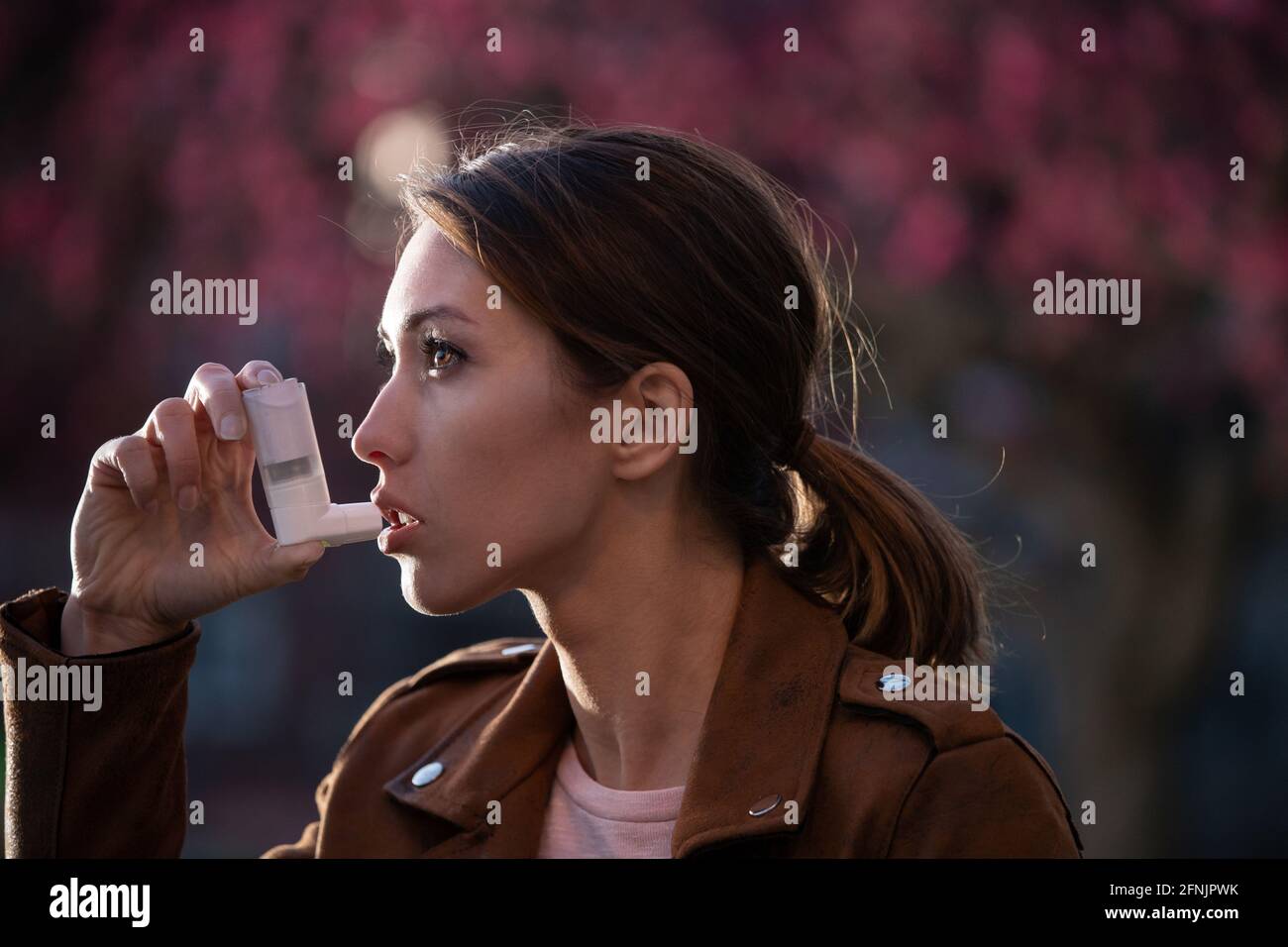 Hübsche junge Frau, die im Frühling vor einem blühenden Baum eine Asthmapumpe benutzt. Saisonaler Allergieanfall Stockfoto