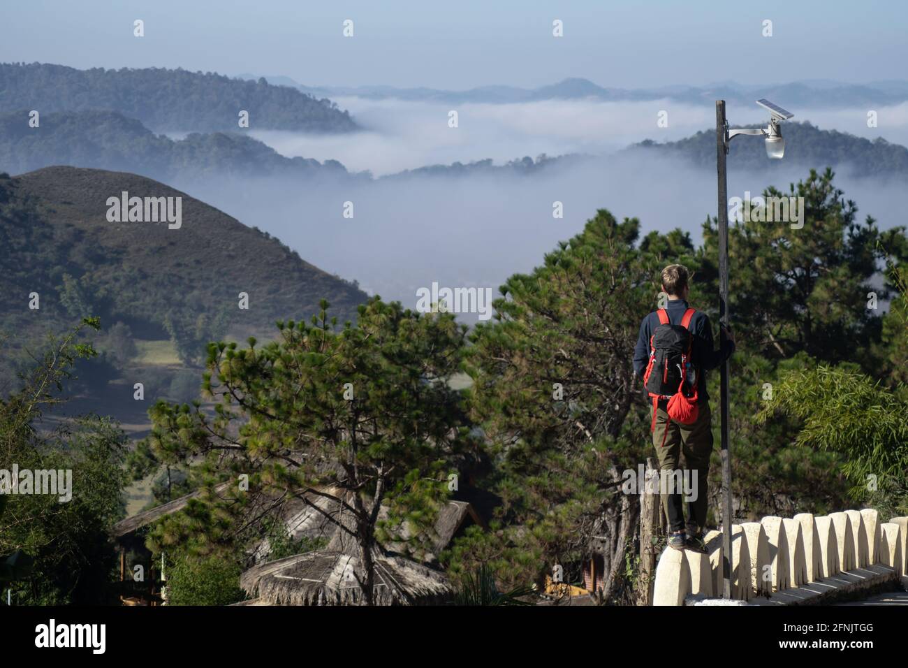 Ein männlicher Toursit mit rotem Rucksack steht auf einem Zaun neben einem Dorf und blickt auf eine neblige Tallandschaft, Shan State, Myanmar Stockfoto