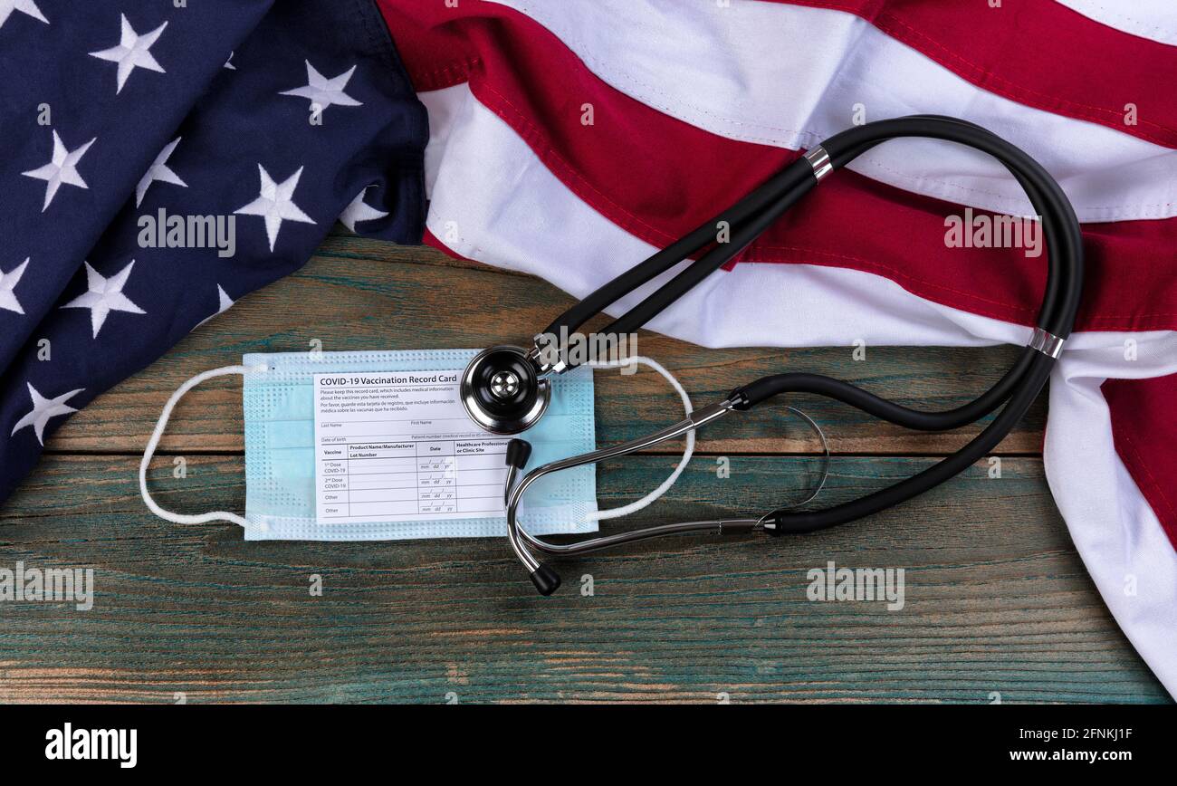 Amerikanische Flagge mit covid 19 Impfpass, persönlicher Gesichtsmask und Stethoskop auf hellblauen rustikalen Holzplanken Stockfoto