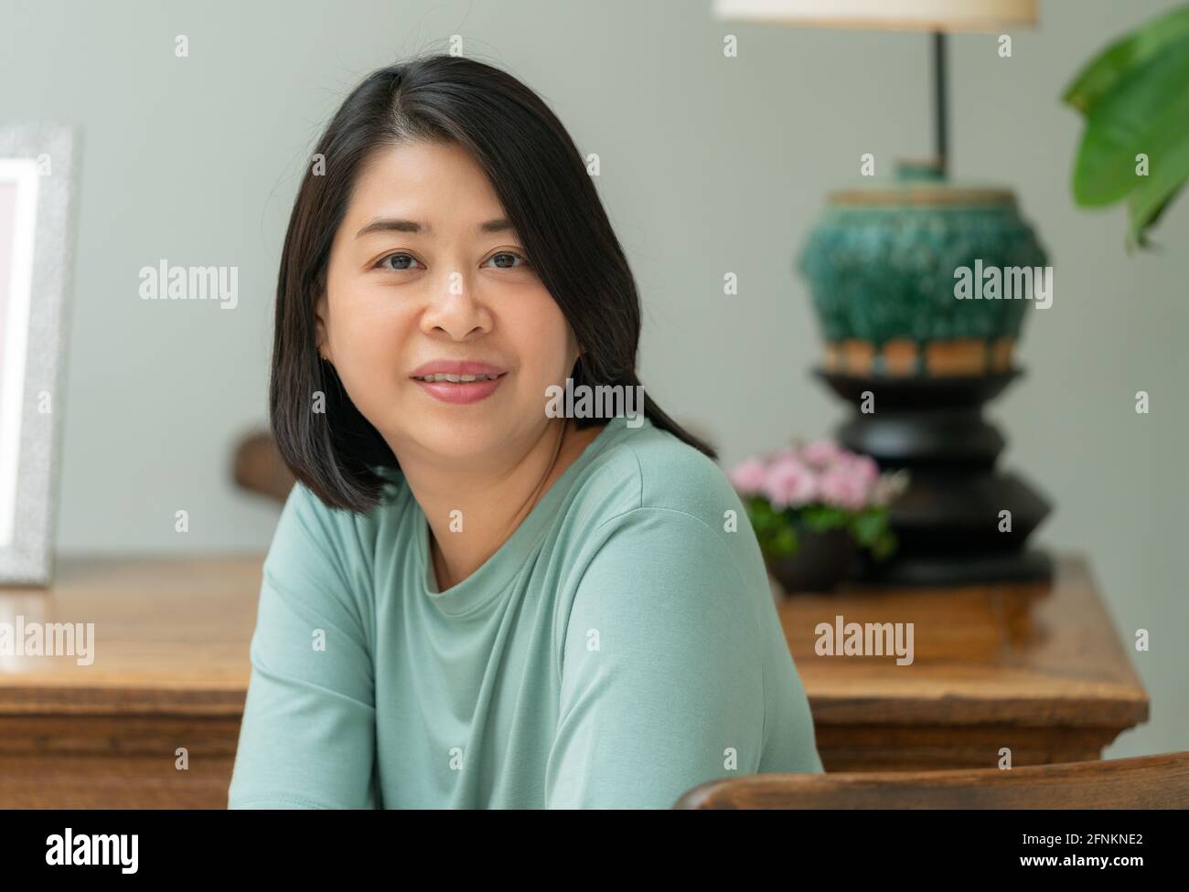 Porträt einer asiatischen Frau mittleren Alters. Sitzen und auf die Kamera schauen, am Holztisch sitzen. Stockfoto