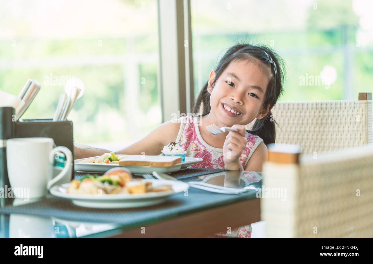 Bezauberndes asiatisches kleines Mädchen, das am Tisch im Hotelrestaurant frühstückte, lächelte und die Kamera betrachte, ein warmes Tonbild. Stockfoto