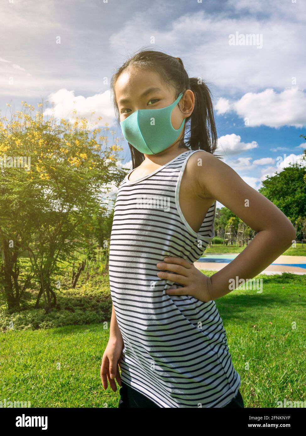 Portrait Asiatische kleine Mädchen posiert im Park mit Arm Akimbo, trägt Schutzmaske für Corona-Virus-Epidemie, Hintergrund blauen Himmel und Blumenbäume, ve Stockfoto