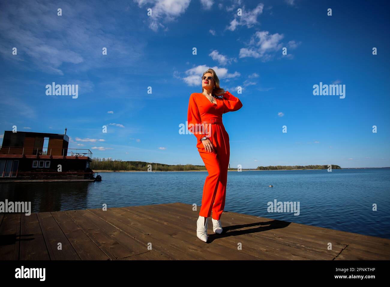 Eine wunderschöne Blondine in Sonnenbrillen und ein roter Anzug vor dem Hintergrund des Sees und des blauen Himmels. Hochwertige Fotos Stockfoto