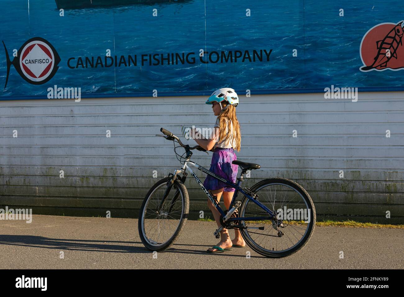 Ein kleines Mädchen mit Helm hielt ihr Fahrrad an, um auf der Straße von Guildford, BC, Kanada, eine Gesichtsmaske zu tragen. April 18,2021. Blick auf die Straße, Reisefoto, Con Stockfoto