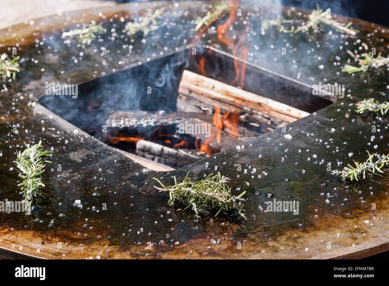 Frische Rosmarinzweige um ein Barbecue-Feuer mit brennendem Feuer Holzstämme und Rauch in einem großen Metallgitter im Freien Stockfoto