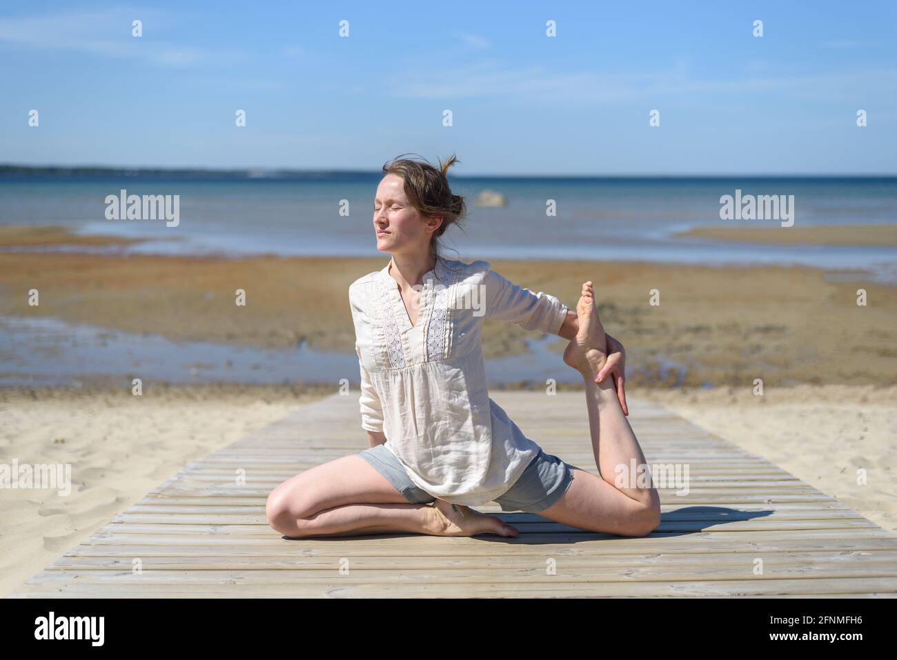 Frau in Leinenkleidung macht Yoga-Stretch-Pose am Strand an sonnigen Tag. Wohlbefinden, körperliche und geistige Gesundheit Stockfoto