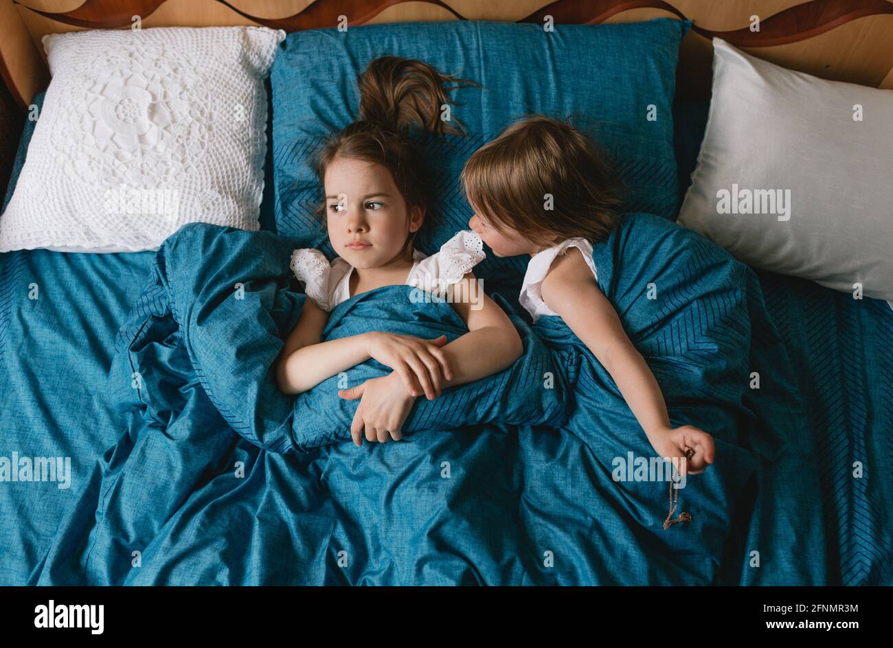 Von oben schauen zwei entzückende Töchter unter der Decke, schauen in die Kamera, liegen zusammen auf dem gemütlichen Bett zu Hause, nette Kinder entspannen sich, genießen Stockfoto