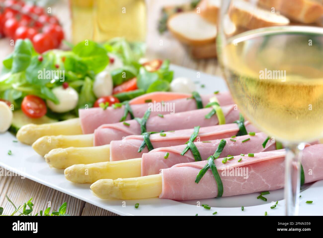 Köstlicher Frühlingssalat mit Spargel, gerollt in gekochtem Schinken auf einer weißen Platte, serviert mit einem jungen trockenen Weißwein und Baguette-Scheiben Stockfoto