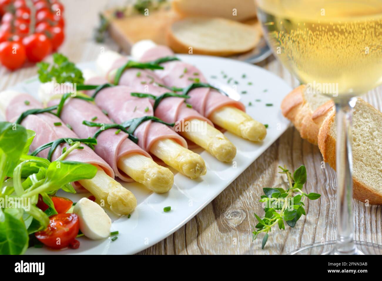 Köstlicher Frühlingssalat mit Spargel, gerollt in gekochtem Schinken auf einer weißen Platte, serviert mit einem jungen trockenen Weißwein und Baguette-Scheiben Stockfoto