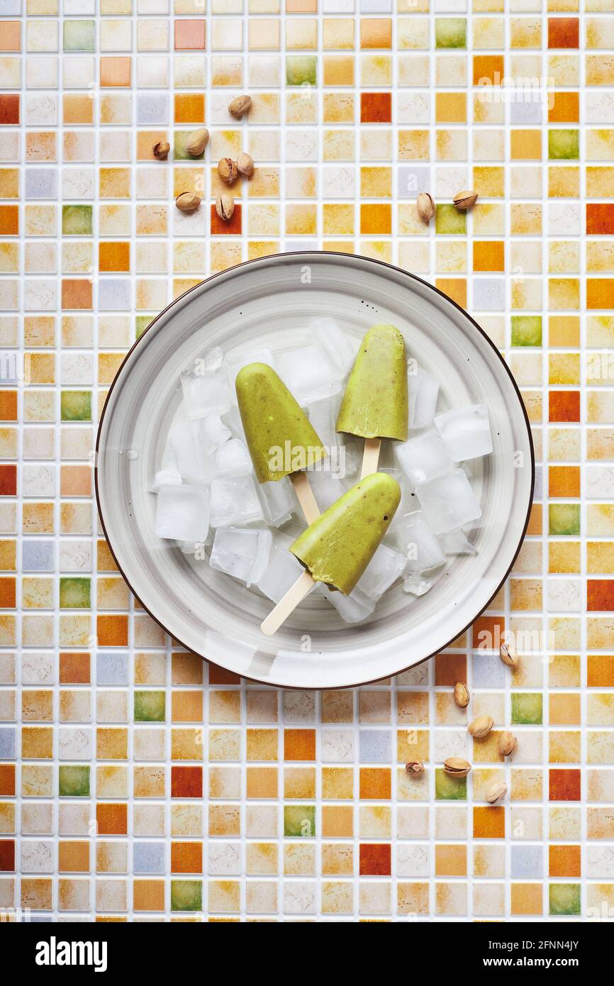 Gefrorener hausgemachter Pistazien-Popsicle in einer Schüssel mit Eis auf einem Tisch mit Mosaikfliesen. Erfrischender Fruchteis, gefrorener grüner Saft auf Stick. Draufsicht, Kopierbereich. Stockfoto