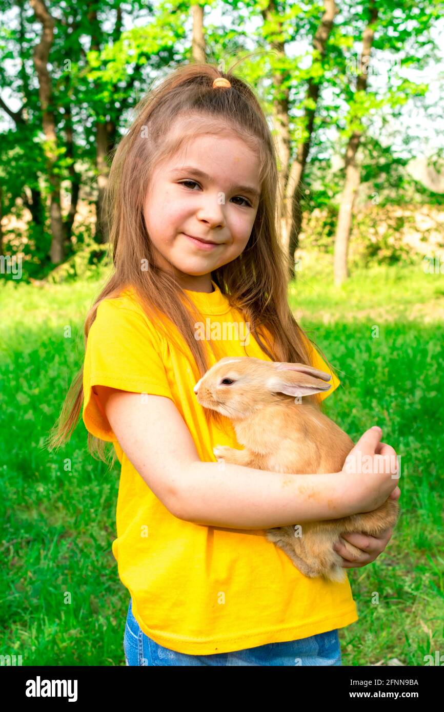 Ein kleines süßes Mädchen in einem gelben T-Shirt hält ein echtes Kaninchen in den Armen vor einem Hintergrund grüner Pflanzen. Im Sommer Aktivitäten im Freien für Kinder Stockfoto