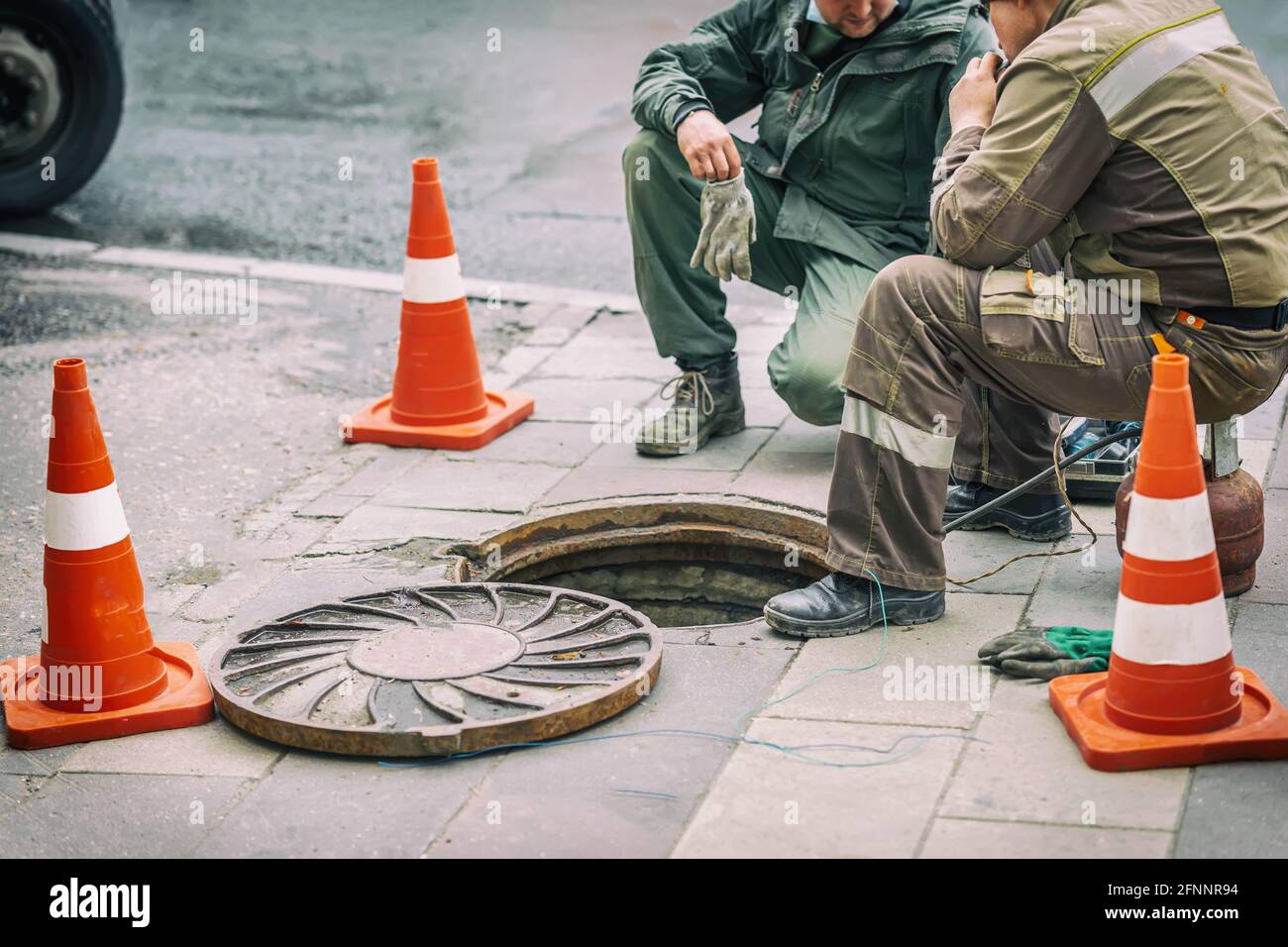 Arbeiter, die auf der Straße in der Nähe von Verkehrskegeln über der offenen Kanalluke sitzen. Reparatur von Abwasser, unterirdisches Versorgungskonzept Stockfoto