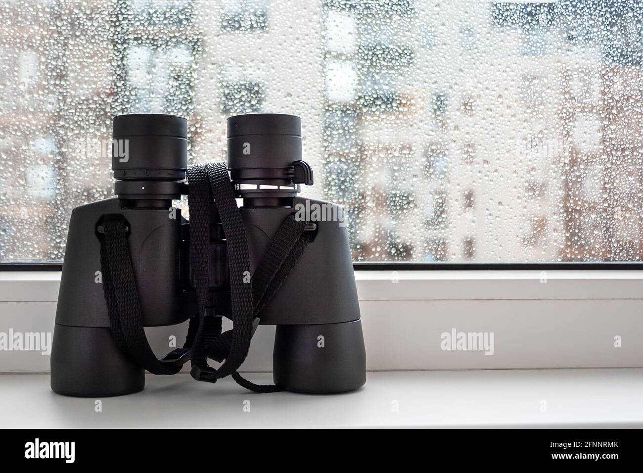 Auf einer weißen Fensterbank am Fenster mit Regentropfen auf dem Glas befindet sich ein schwarzes Fernglas zur Beobachtung von Nachbarn. Faszinierende Beobachtung der Umwelt Stockfoto