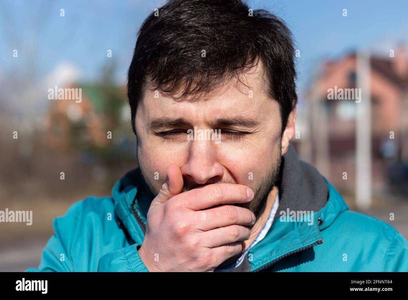 Hustet, niest oder gähnt, kneikender bärtiger Mann, der an einem sonnigen Tag seinen Mund mit der Hand auf der Straße bedeckt. Kranker oder müder Mann. Nahaufnahme. B Stockfoto