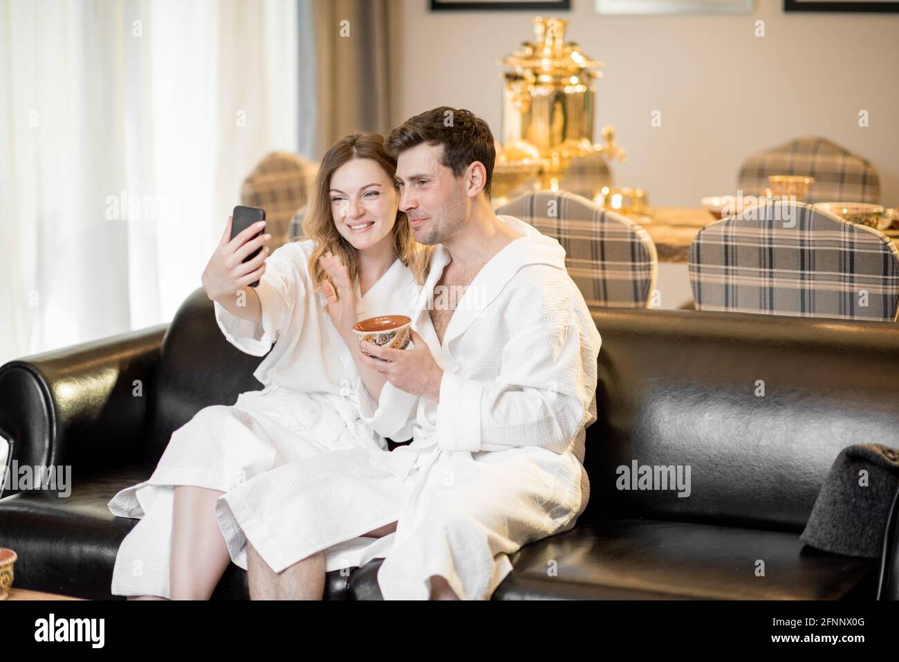 Frau und Mann im Bademantel sitzen auf dem Sofa im Wohnzimmer des Wellnesshauses und machen ein Selfie. Relax und Wellness Konzept. Paare verbringen und genießen Zeit zusammen. Speicherplatz kopieren. Stockfoto