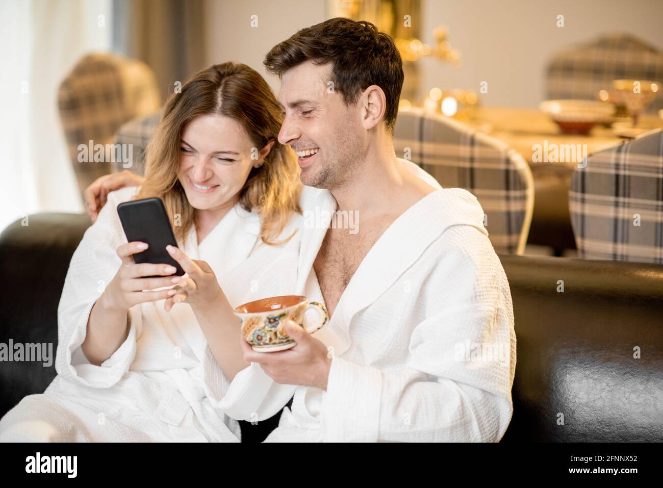 Frau und Mann im Bademantel sitzen auf dem Sofa im Wohnzimmer des Wellnesshauses und machen ein Selfie. Relax und Wellness Konzept. Paare verbringen und genießen Zeit zusammen. Stockfoto