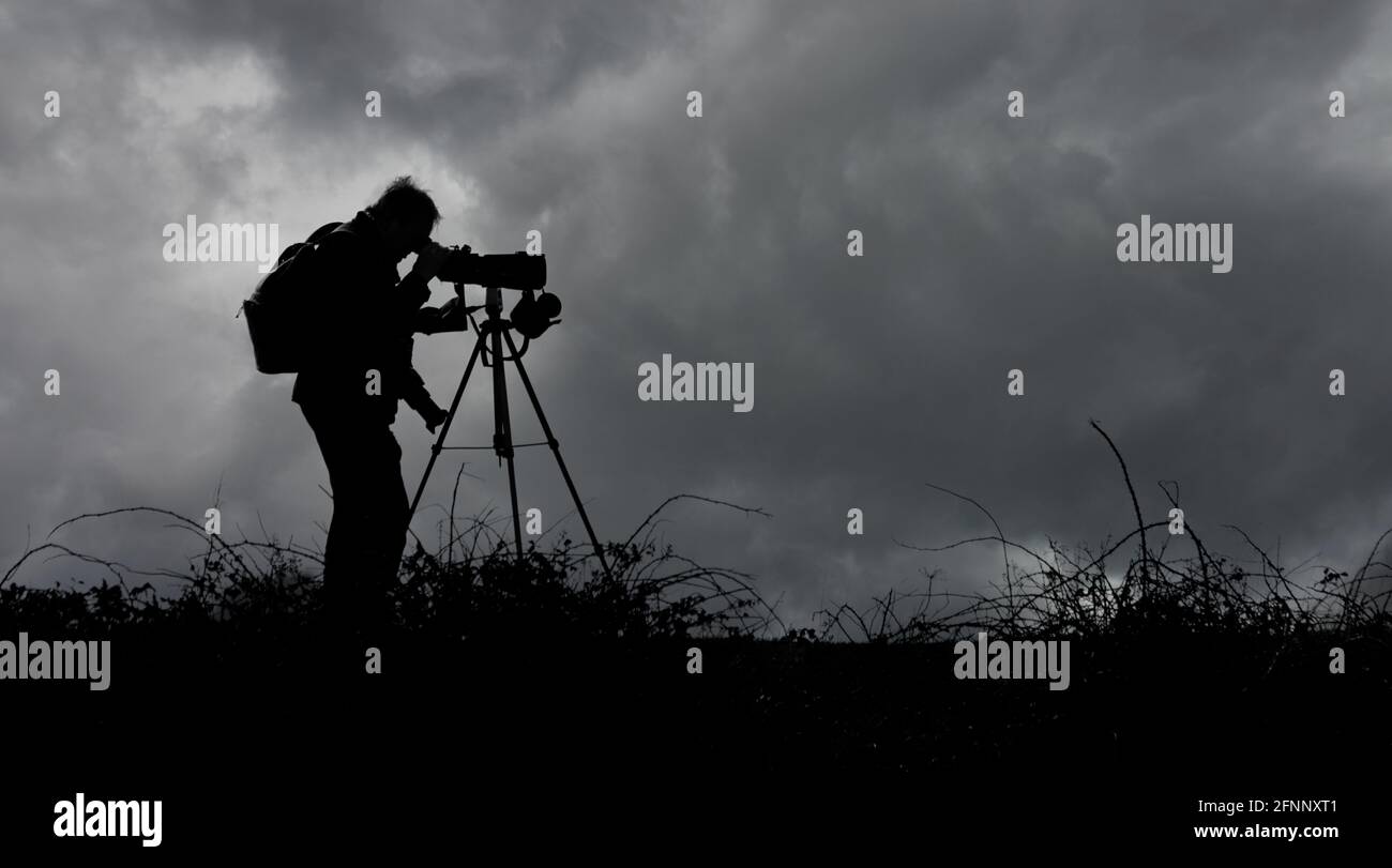 Bird Watcher, Twitcher, Looking Through A Telescope on A Tripod on the Horizon Against A Dark Stormy Sky, Großbritannien. Schwarzweiß, Schwarz und Weiß Stockfoto