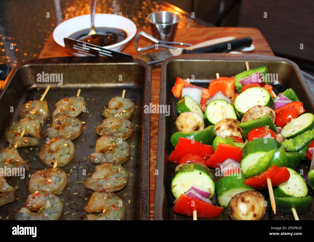 Nahaufnahme von Garnelen und Gemüsespiessen in Pfannen auf einem Schneidebrett, bevor sie gegrillt werden. Grillsoße und Geschirr im Hintergrund nicht im Fokus. Stockfoto