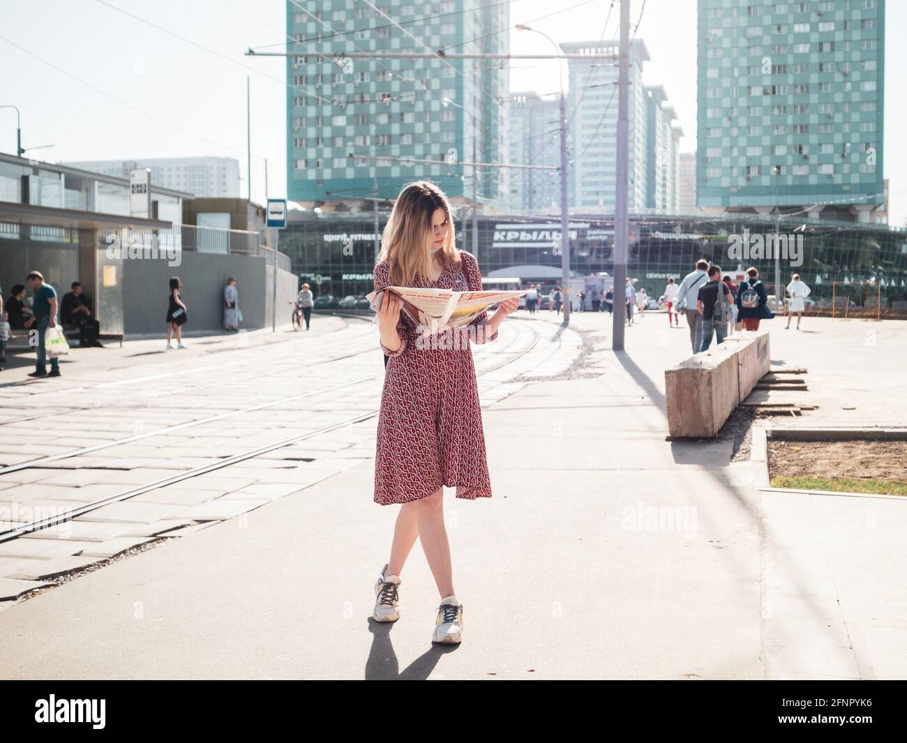 Moskau. Russland. 18.Mai 2021. Ein junges Mädchen in einem hellroten Kleid steht mit gekreuzten Beinen und liest eine Zeitung. Helles Sonnenlicht erhellt die Stadtstraße Stockfoto