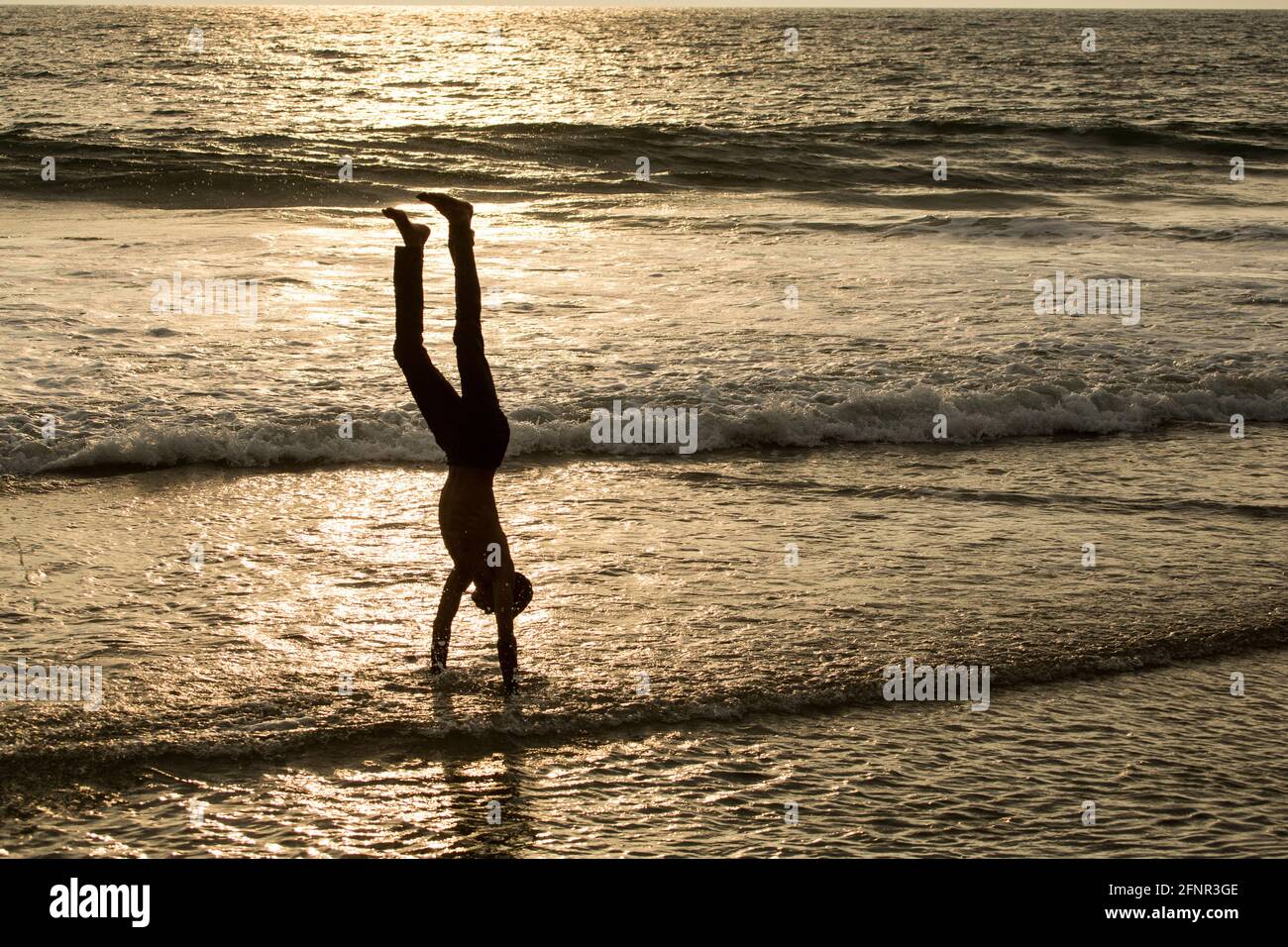 Ein Junge stellt einen Handstand in den Wellen des Meeres und kann als Silhouette vor dem Licht des Sonnenuntergangs gesehen werden, der sich in den Wellen widerspiegelt. Stockfoto