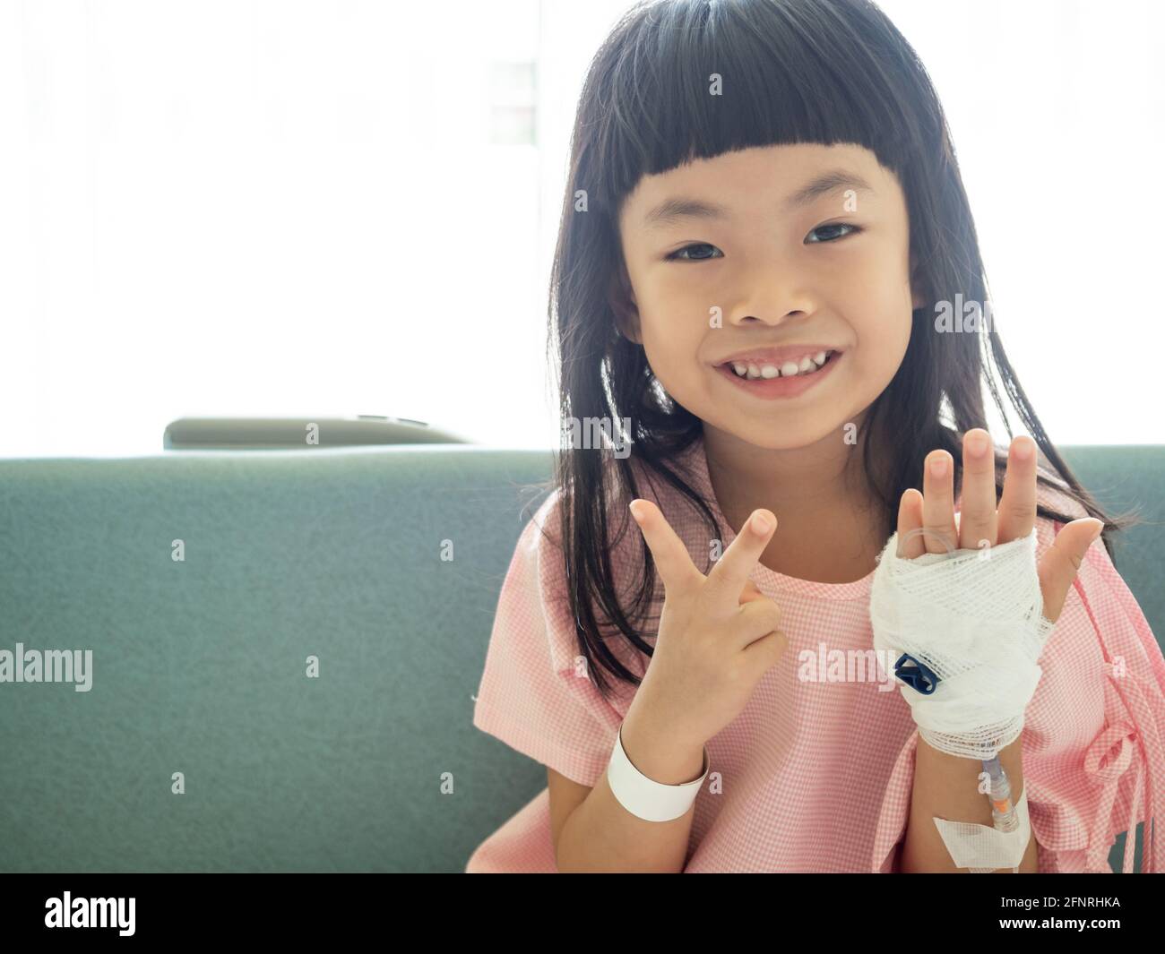 Asiatische Kind Mädchen sitzt auf Krankenhausbett, zeigt zwei Finger. Positives Bild des Patienten im Krankenhaus. Stockfoto