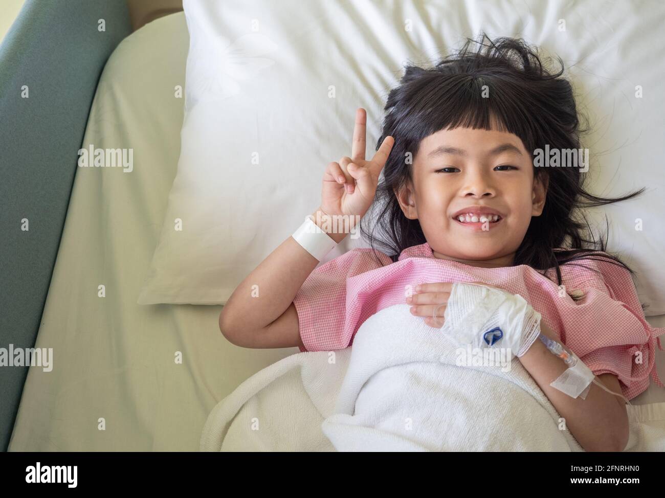 Asiatisches Mädchen zeigt zwei Finger auf dem Krankenhausbett. Positive Empfindung eines geduldigen Kindes. Stockfoto