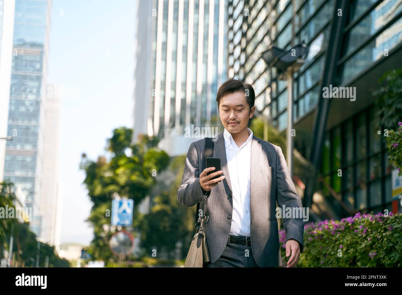 asiatischer Geschäftsmann, der beim Gehen zur Arbeit auf das Mobiltelefon schaut Im zentralen geschäftsviertel der modernen Stadt Stockfoto