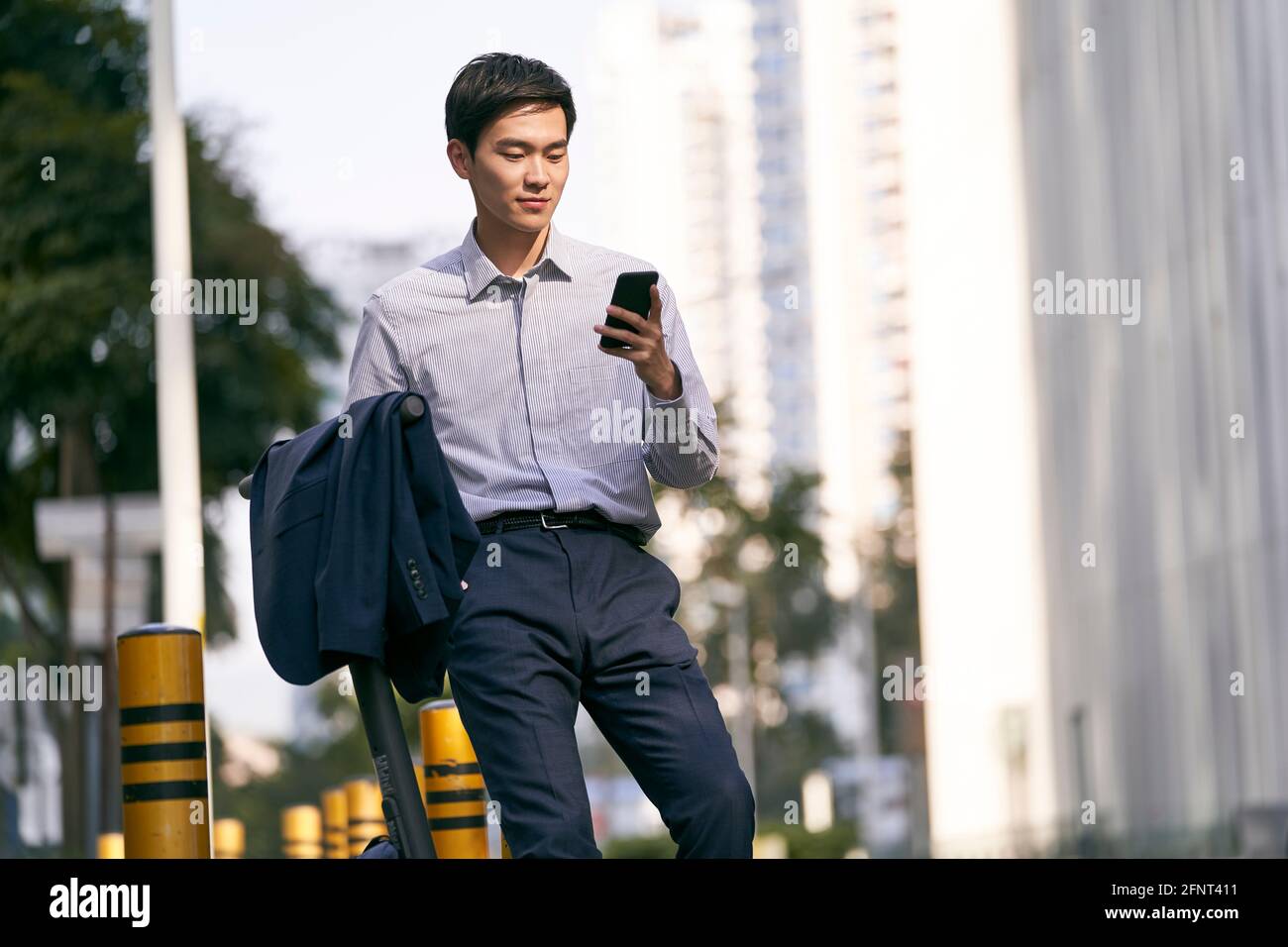 Junger asiatischer Geschäftsmann, der neben seinem Elektroroller steht und sucht Am Handy auf der Straße im Abtrieb der modernen Stadt Stockfoto