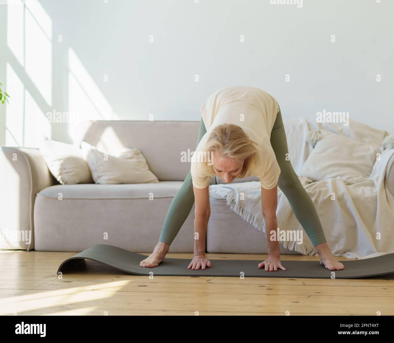 Aktive ältere 60s Frau im Sport tragen nach unten schauende Hund Yoga-Pose üben, während Sie zu Hause trainieren. Reife flexible Dame tun breitbeinigen Vorwärtsbeuge oder Prasarita Padottanasana Haltung Stockfoto