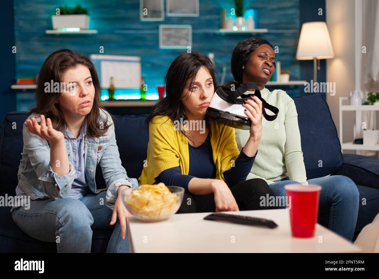 Traurige Frauen, die nach dem Verlust des Virtual-Reality-Gaming-Wettbewerbs das Headset auf der Couch halten. Gemischte Renngruppe von Menschen, die sich zusammen hingen und sich spät in der Nacht im Wohnzimmer amüsieren Stockfoto