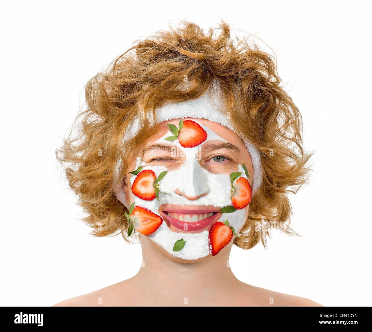 Rothaarige lockige junge Frau mit einer positiven fröhlichen Stimmung, lächelnd und zwinkert, geschlossenes Auge. Mit einer kosmetischen Maske auf dem Gesicht von Erdbeeren und Creme. Stockfoto