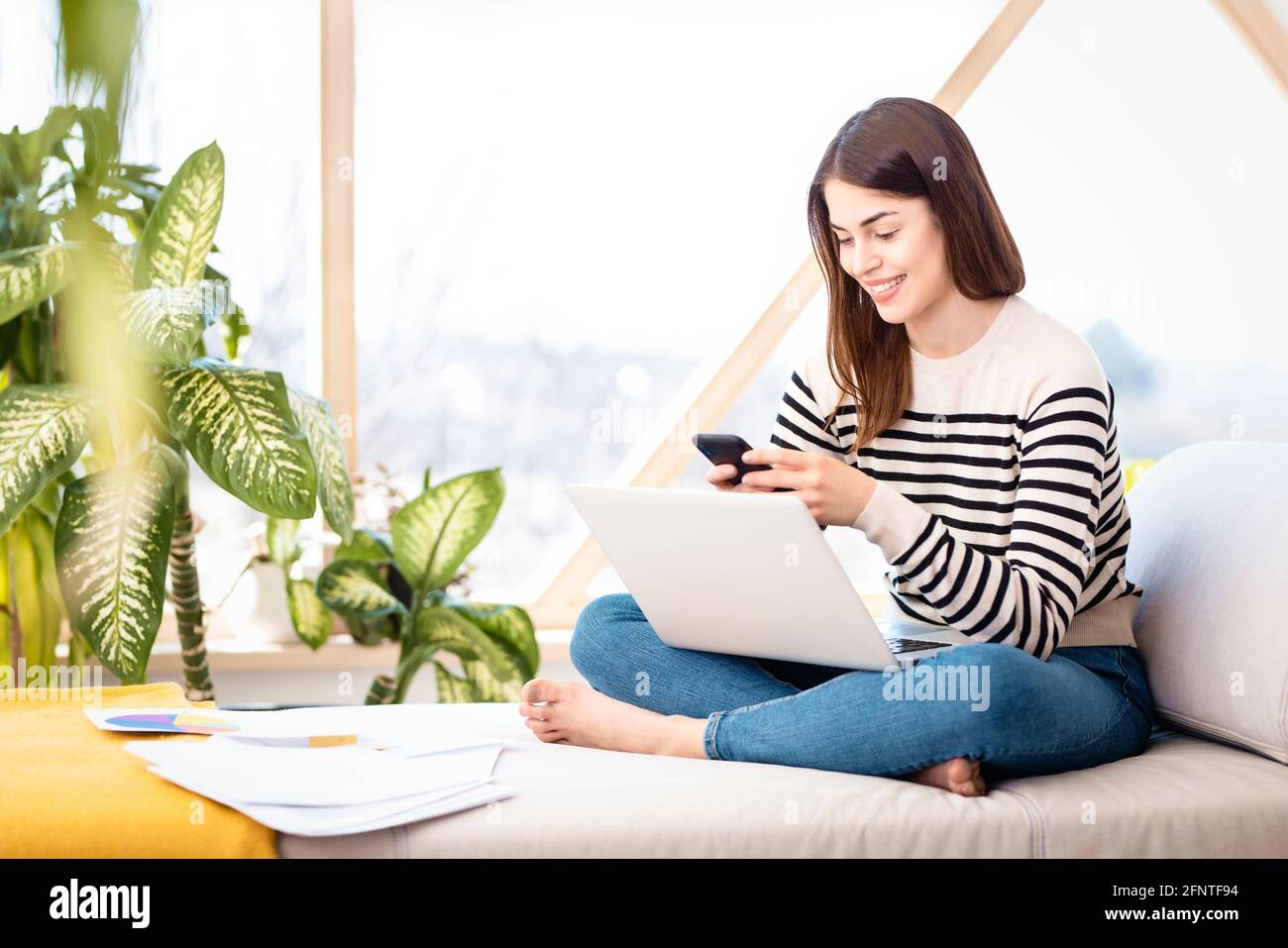 Aufnahme einer jungen Frau, die ihren Laptop und SMS verwendet, während sie von zu Hause aus arbeitet. Heimbüro. Stockfoto