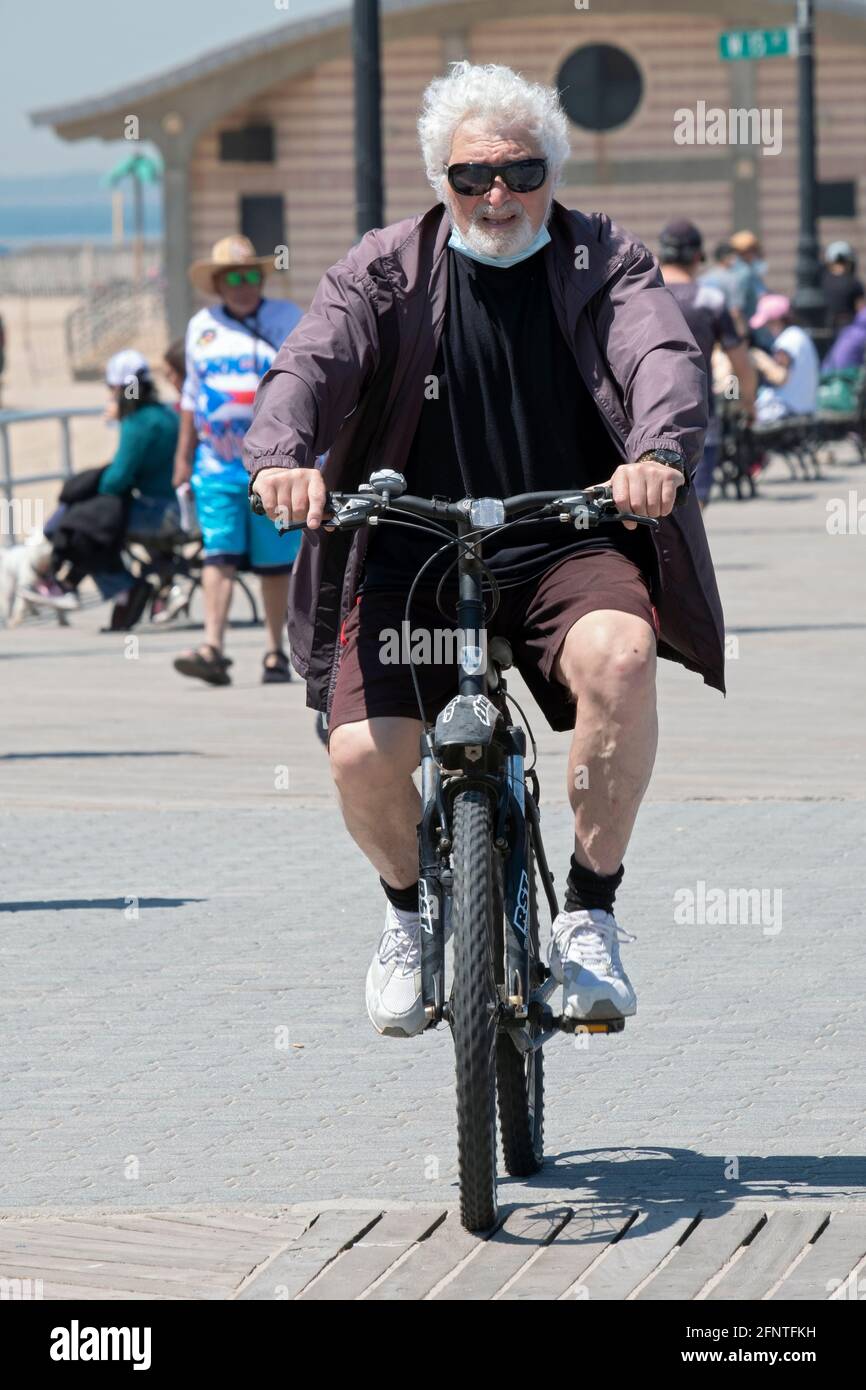 Ein älterer Mann mit weißen Haaren und einem Spitzbart fährt auf dem Boardwalk in Coney Island, Brooklyn, New York City, mit dem Fahrrad. Stockfoto