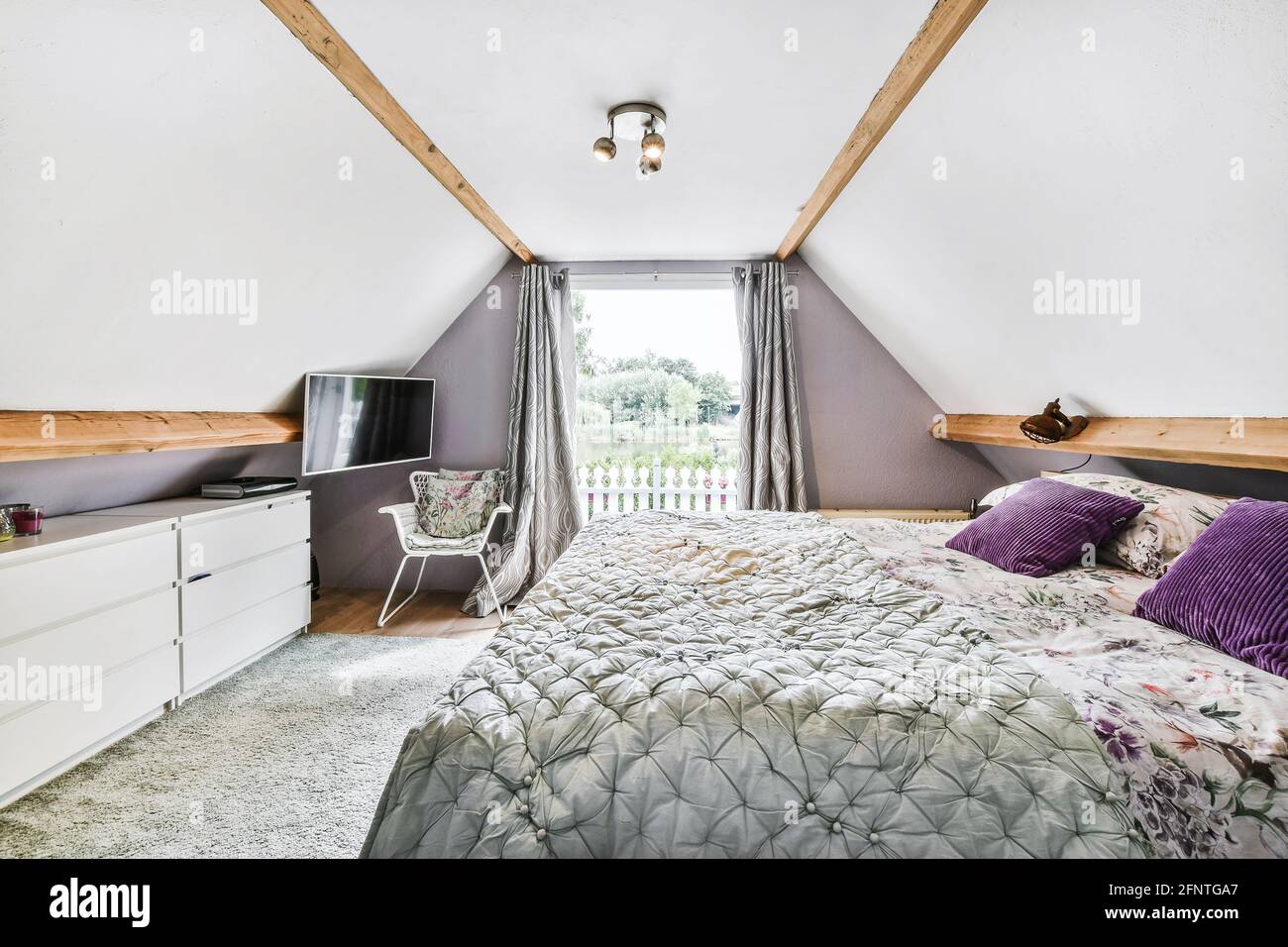 Komfortables Bett mit Kissen und Decke neben Stuhl und Fernseher gegen  Fenster mit Vorhängen im hellen Schlafzimmer unter dem Dach Zu Hause  Stockfotografie - Alamy