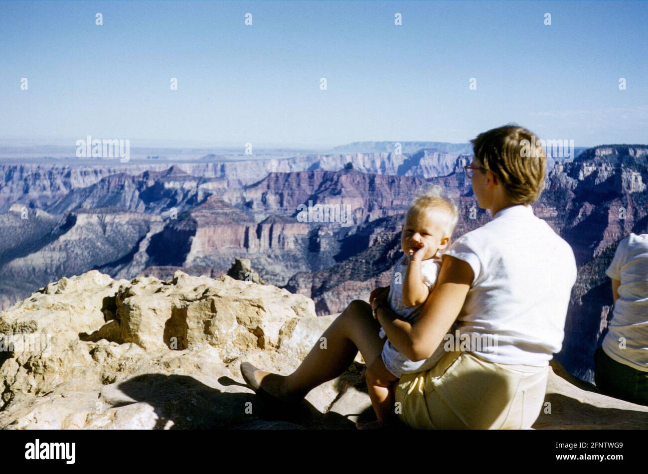 Junge Frau in Shorts mit einem 7 bis 8 Monate alten blonden Baby, das in den 1950er Jahren auf einem Aussichtspunkt auf einer Klippe mit Blick auf den Grand Canyon, Colorado, USA sitzt Stockfoto