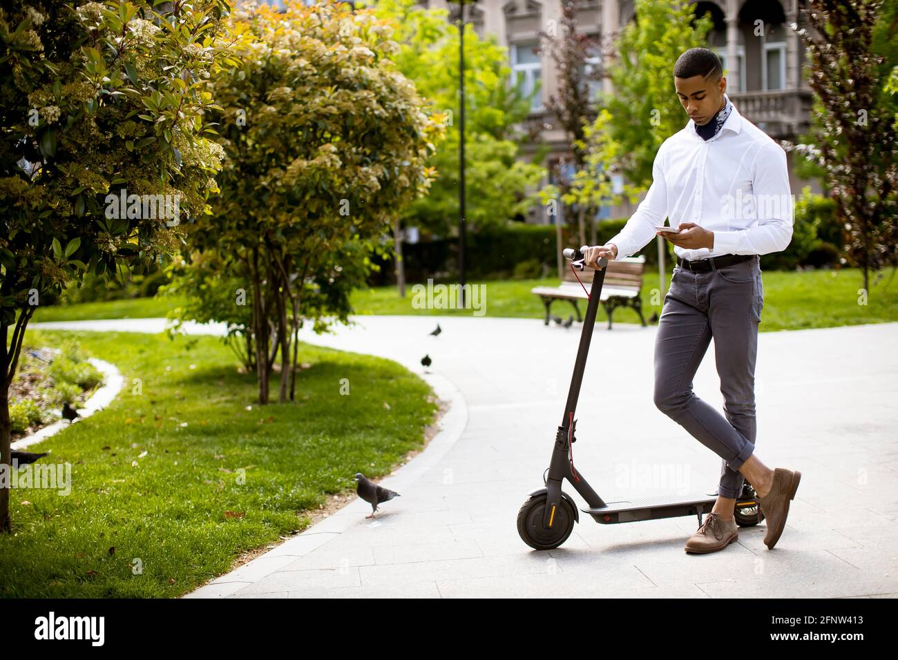 Hübscher junger Afroamerikaner, der im Stehen ein Mobiltelefon benutzt Elektroroller auf einer Straße Stockfoto