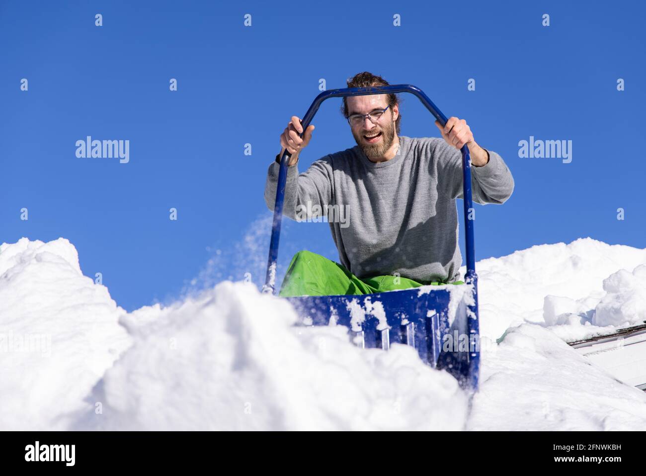 Von unten geschossen eines jungen Mannes, der mit einer blauen Handschaufel vom Dach eines Hauses posiert, wo er an einem sonnigen Wintertag frischen Schnee entfernt. Stockfoto