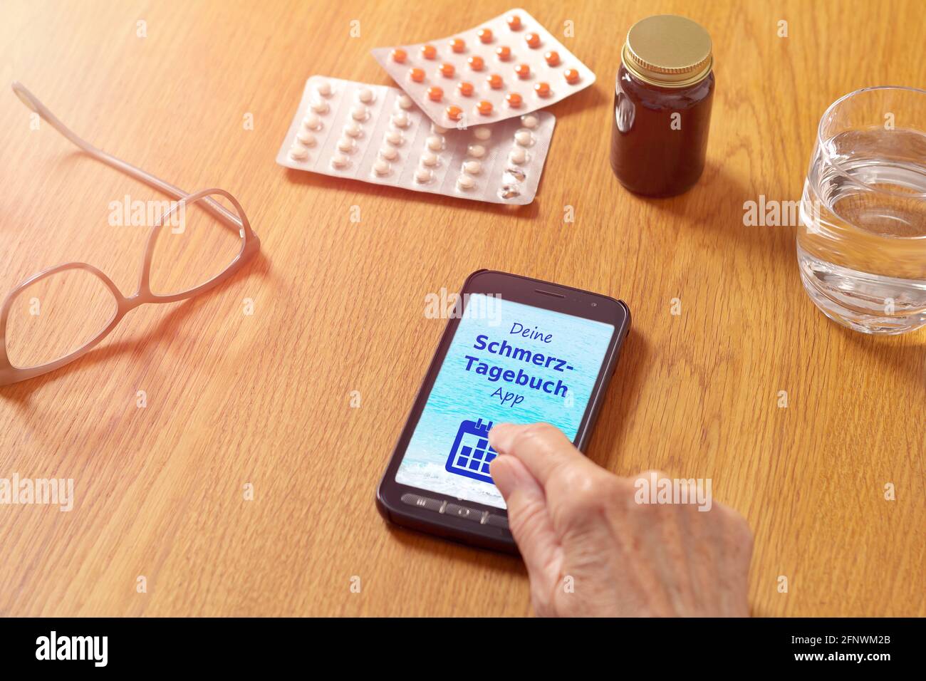 Medizin auf einem Tisch und eine Hand berühren ein Smartphone mit einer deutschen mobilen Anwendung, Text: Schmerztagebuch App. Übersetzung: Ihre Schmerztagebuch App. Stockfoto