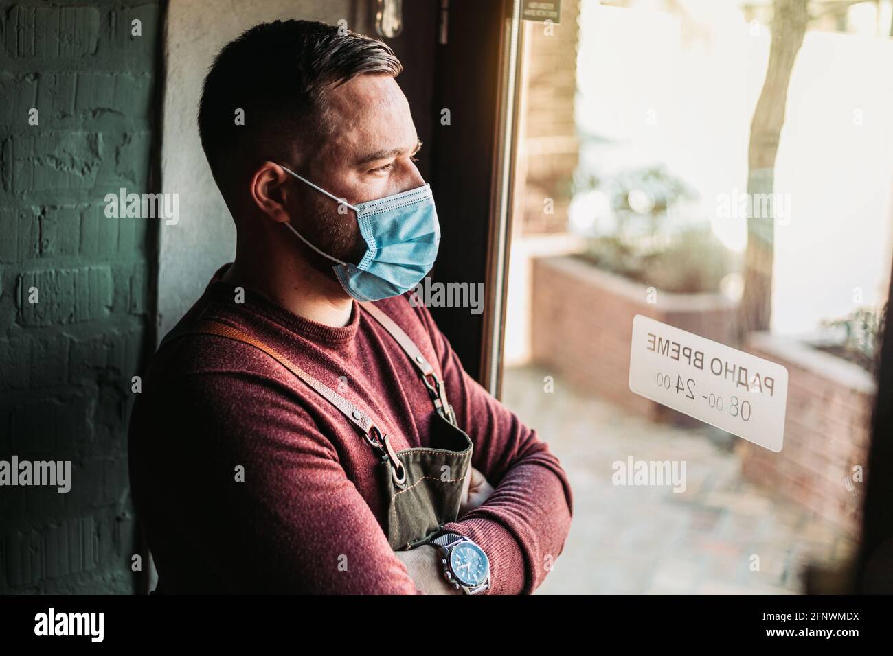 Junger Kaffeehaus, der am Eingang stand und aufgeregt aussehte. An der Tür steht die Arbeitszeit in serbischer Sprache Stockfoto