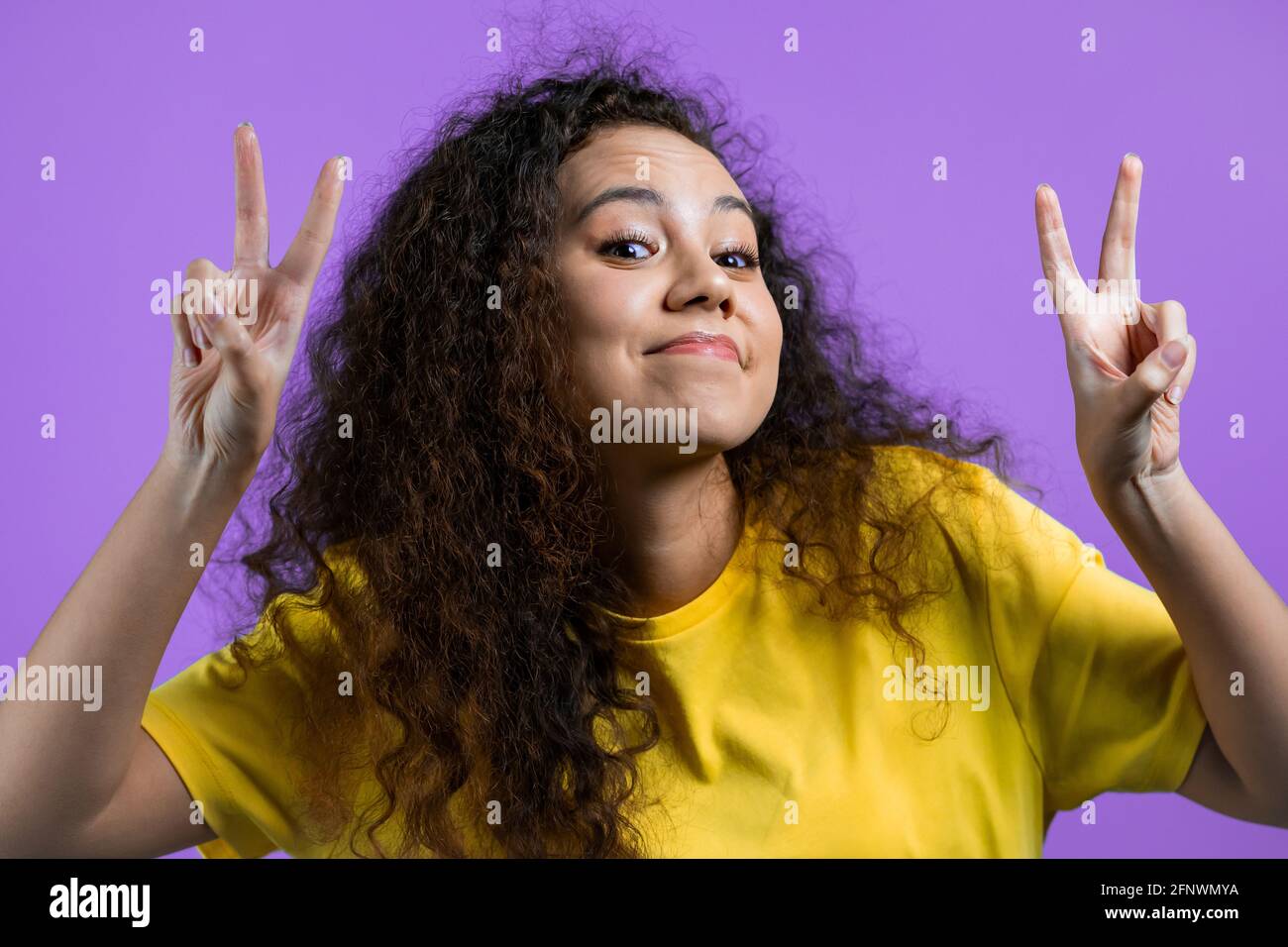 Hübsche Frau mit lockigen Haaren zeigt mit Händen und zwei Fingern Luft Zitate Geste, biegen Finger isoliert über violetten Hintergrund. Nicht lustig, Ironie und Stockfoto