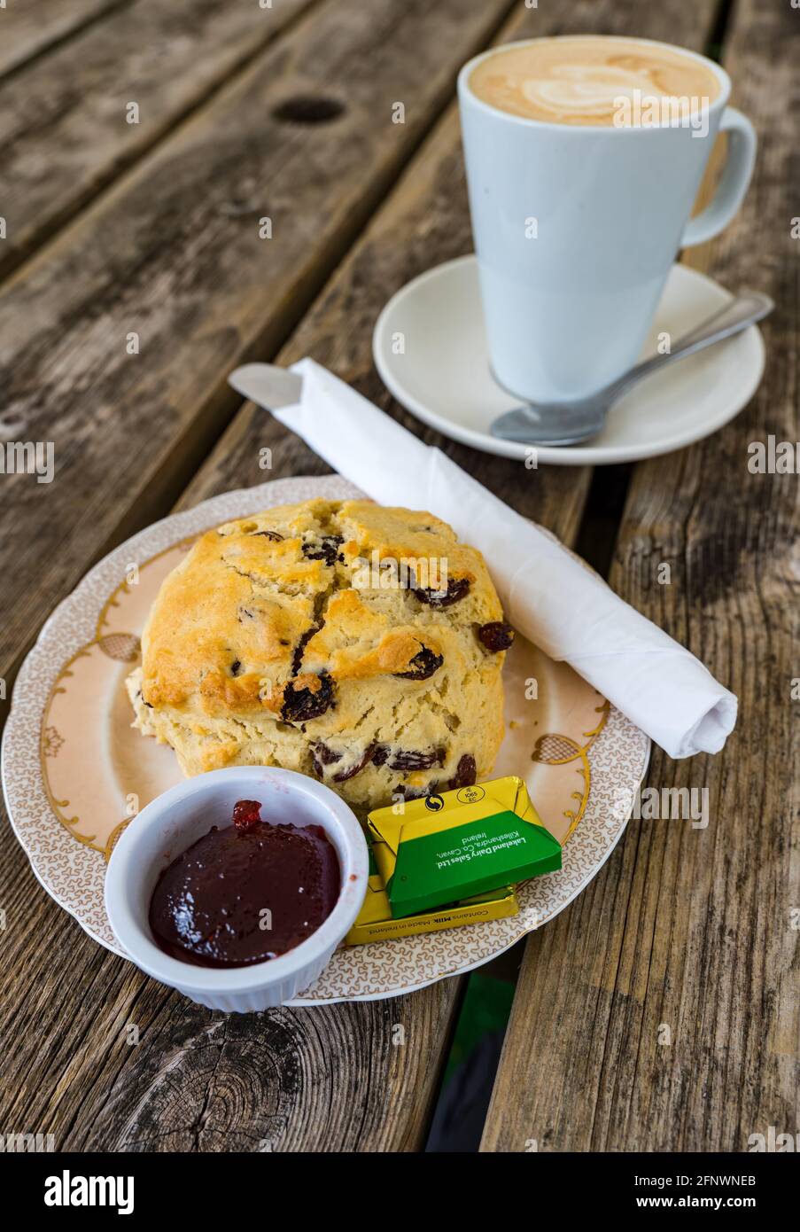 Hausgemachter Fruchtkegel mit Marmelade und Butter auf dem Teller serviert Auf dem Kaffeetisch im Freien mit einem Latte-Becher Kaffee Stockfoto