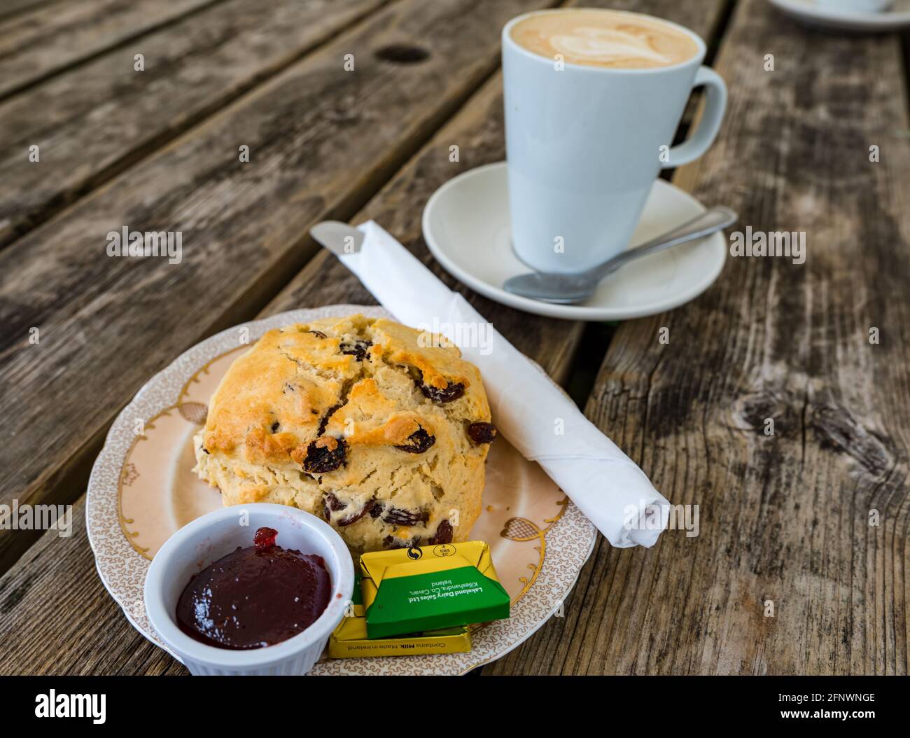 Hausgemachter Fruchtkegel mit Marmelade und Butter auf dem Teller serviert Auf dem Kaffeetisch im Freien mit einem Latte-Becher Kaffee Stockfoto