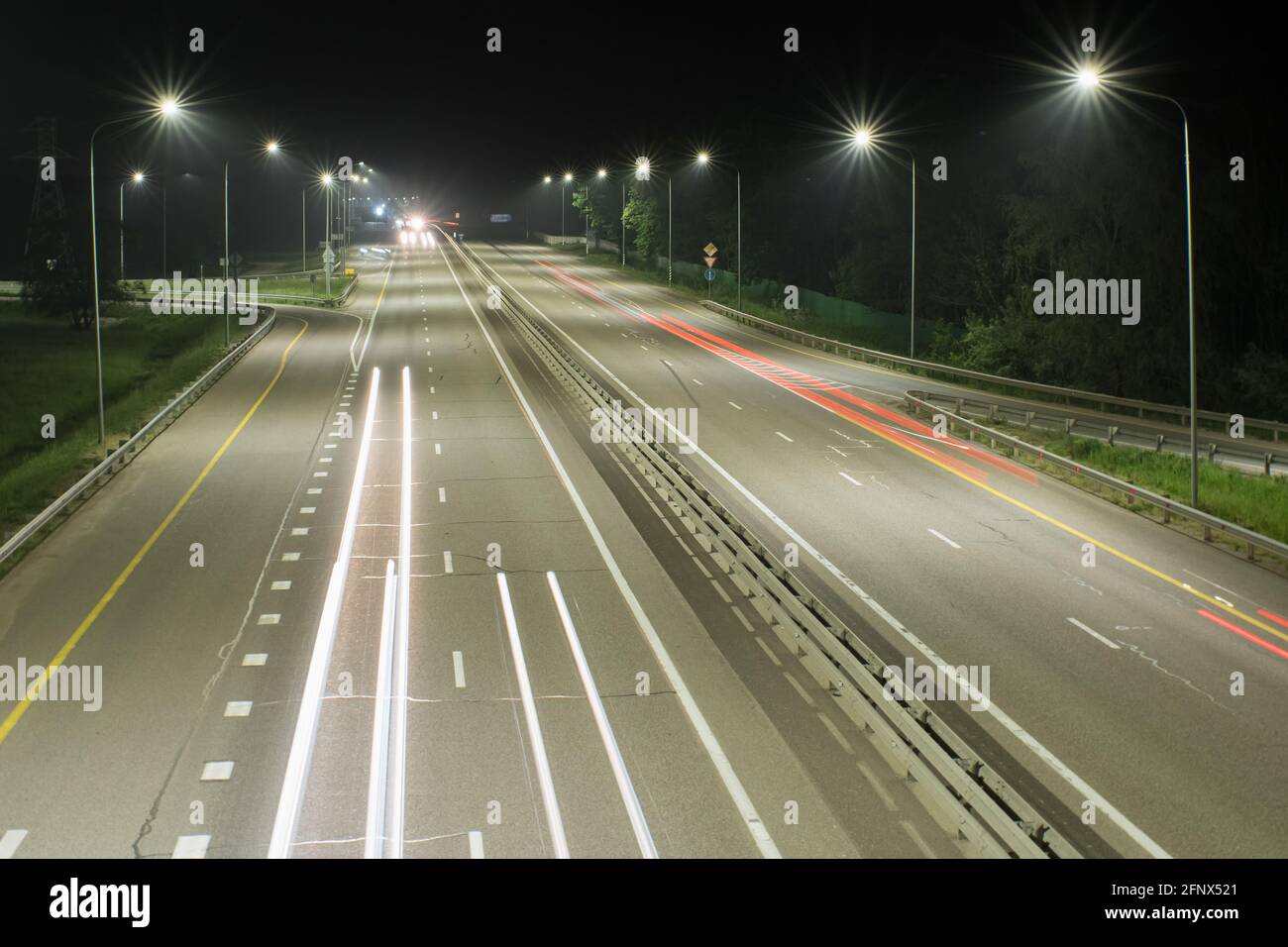 Nachtbahn, beleuchtet von Laternen. Es zeigt Lichtstreifen aus den Scheinwerfern vorbeifahrener Autos. Nachtszenen Stockfoto