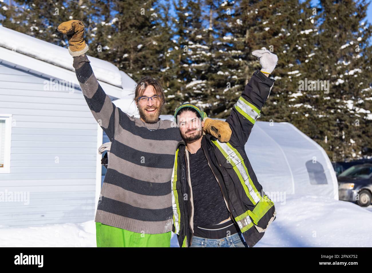 Porträt zweier lächelnder, gutwilliger junger Männer, die nach einem harten Tag des Schnees in einem kanadischen Landhaus mit den Armen nach oben für die Kamera posieren. Stockfoto