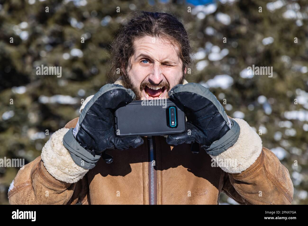 Dramatisch ironisches Porträt eines jungen Mannes in einem Wintermantel, der mit seinem Mobiltelefon ein Foto macht und dabei ein übertriebenes, witziges Gesicht macht. Stockfoto