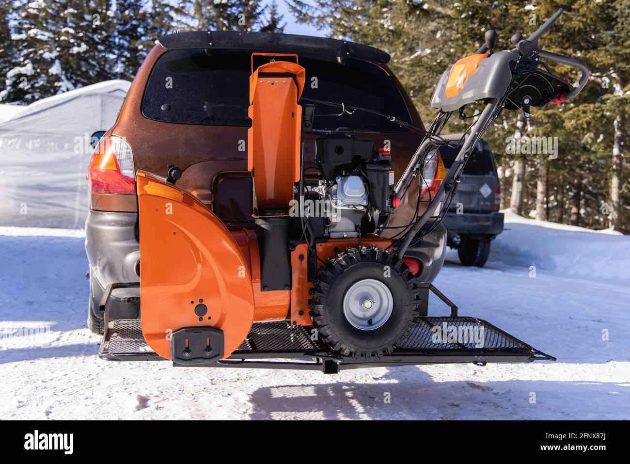 Ein leuchtend orangefarbener mechanischer Schneepflug wird vor einem Auto in einer Auffahrt geparkt, der bereit ist, in einem kanadischen Landhaus Schnee zu entfernen. Stockfoto
