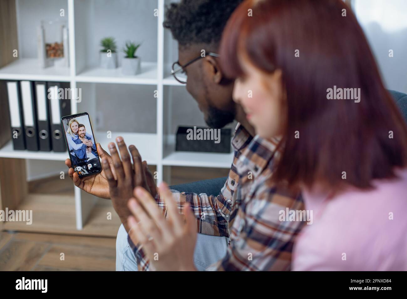 Rückansicht eines multiethnischen Paares, das während eines Videogesprächs mit Freunden auf dem Smartphone die Hände winkt. Menschen, die moderne Technologie nutzen, während sie zu Hause bleiben. Stockfoto