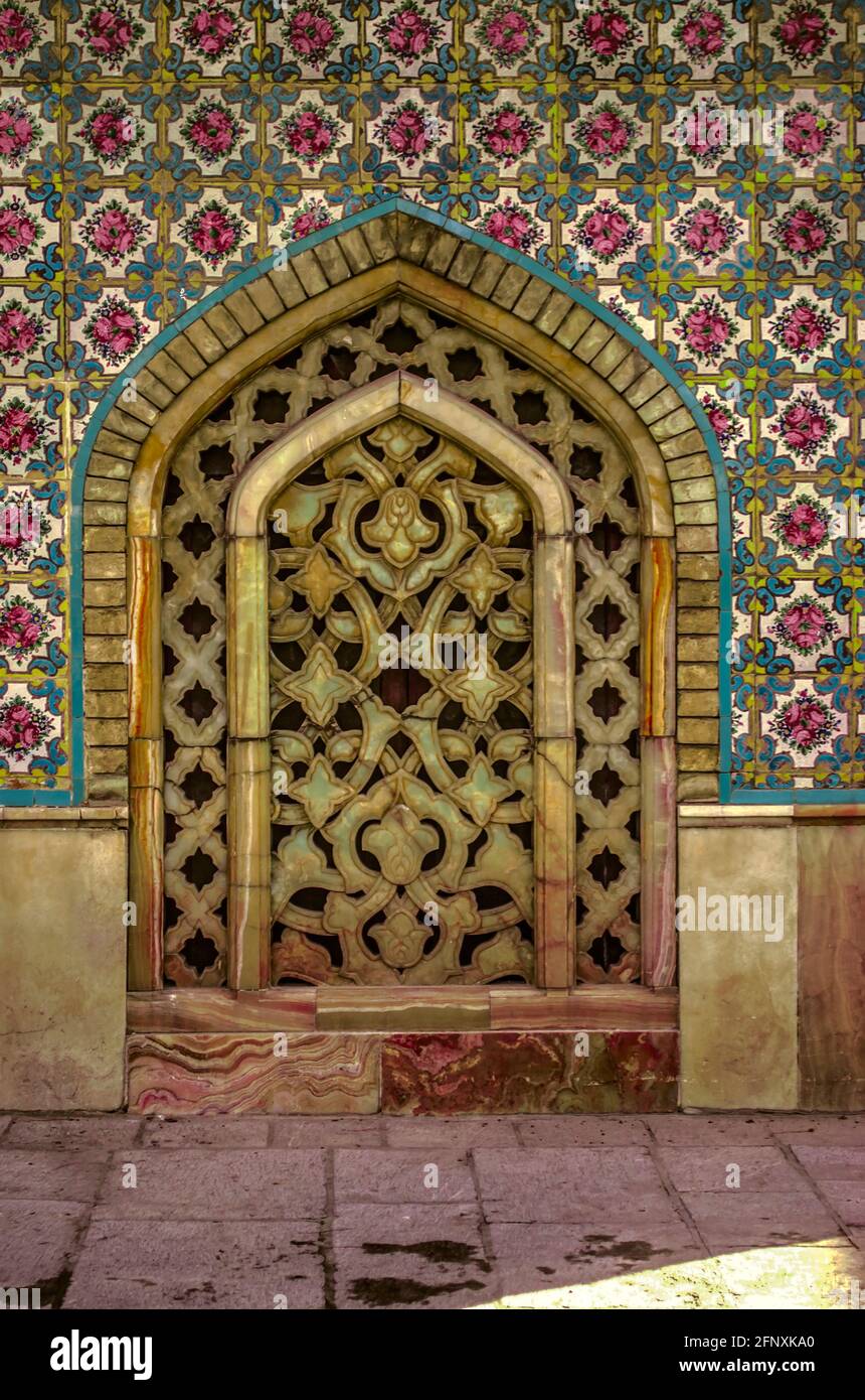Wand mit einem Muster aus Keramikfliesen mit einem ovalen Loch, das mit einem dekorativen Gitter aus geschnitztem gelbem und rosafarbenem Marmor im Golestan-Palast bedeckt ist. Stockfoto