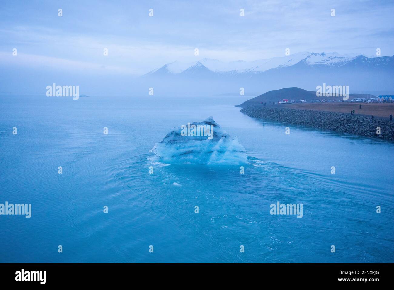 Isländischer Gletscher, der durch die Auswirkungen der globalen Erwärmung schwimmt. Neblige Atmosphäre mit schneebedeckter Bergkette in der Ferne. Eisberg hat Schichten von Vulkanen Stockfoto